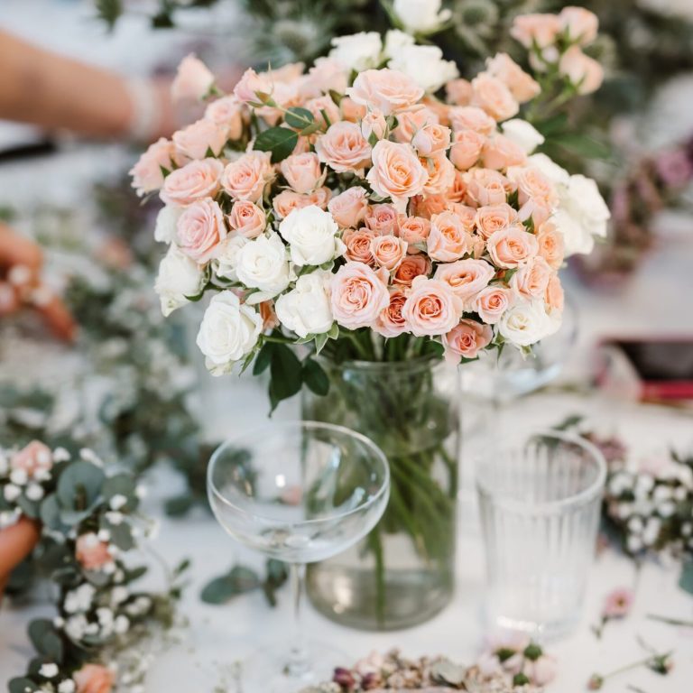 Rosen in weiß und rosa stehen auf dem Tisch bei einem JGA an dem Blumenkränze im Hintergrund gebunden werden.