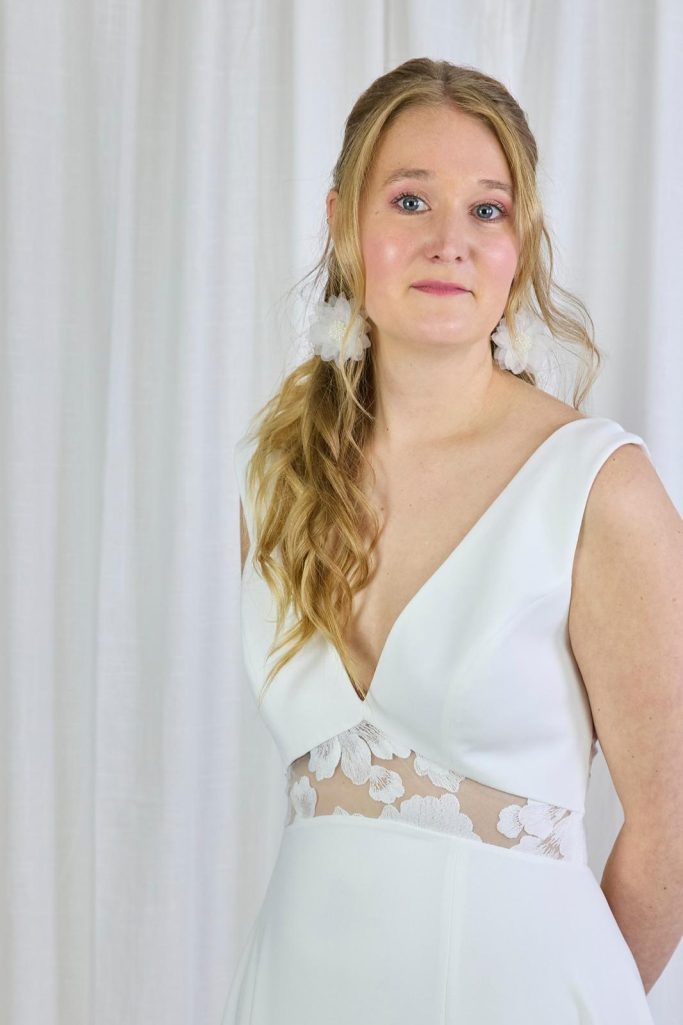 LAMBERT CREATIONS Braut schaut lächelnd mit einem weißen Brautkleid und großen Blumenohrringen in die Kamera.