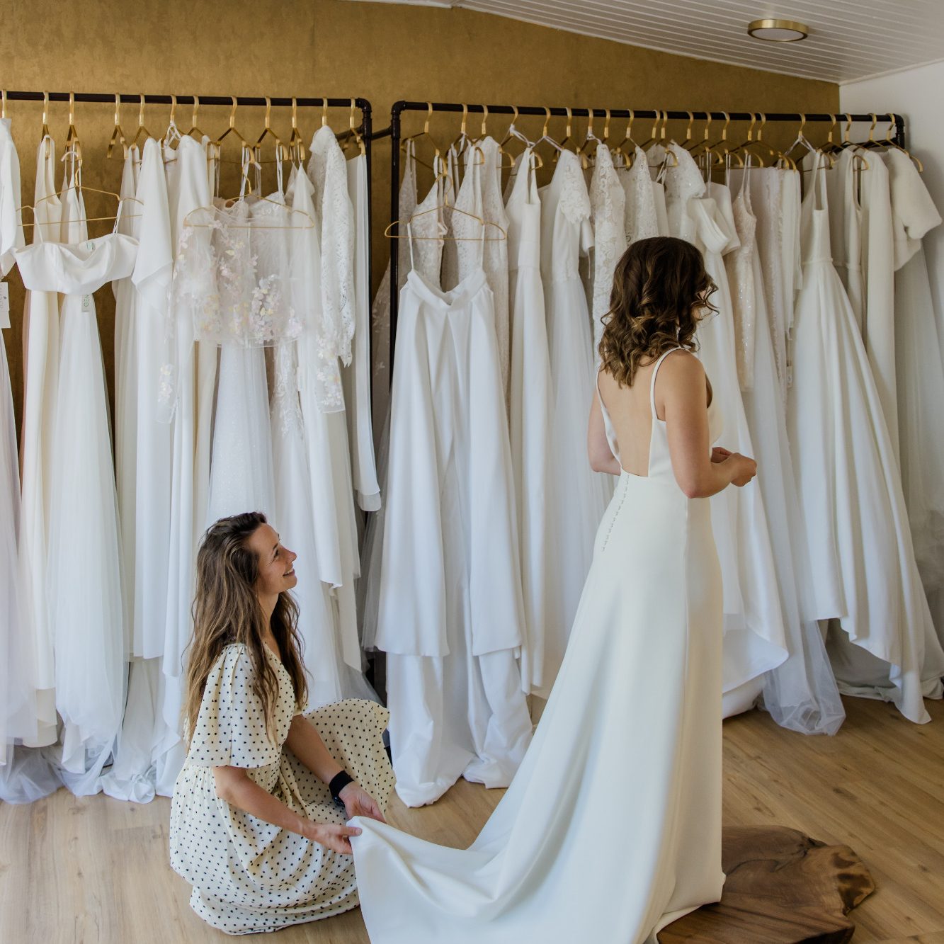 Brautkleid Anprobe in Kiel spontan möglich. Eine Braut steht mit ihrem Brautkleid auf einem Holzpodest vor einer Stange mit Brautkleidern.