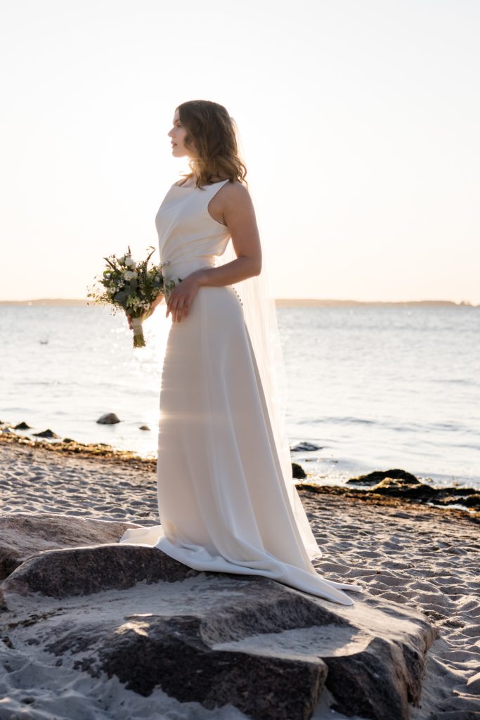 Clean Bridal Look aus Stretch-Crepe wird am Strand an der Ostsee getragen. Die Braut hält einen Brautstrauß in der Hand und steht im Sand.