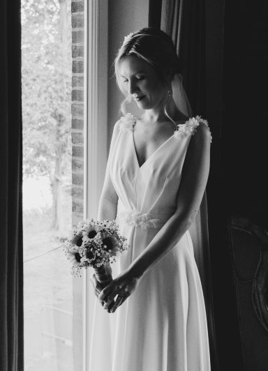Braut schaut Brautstrauß an und steht verträumt am Fenster.