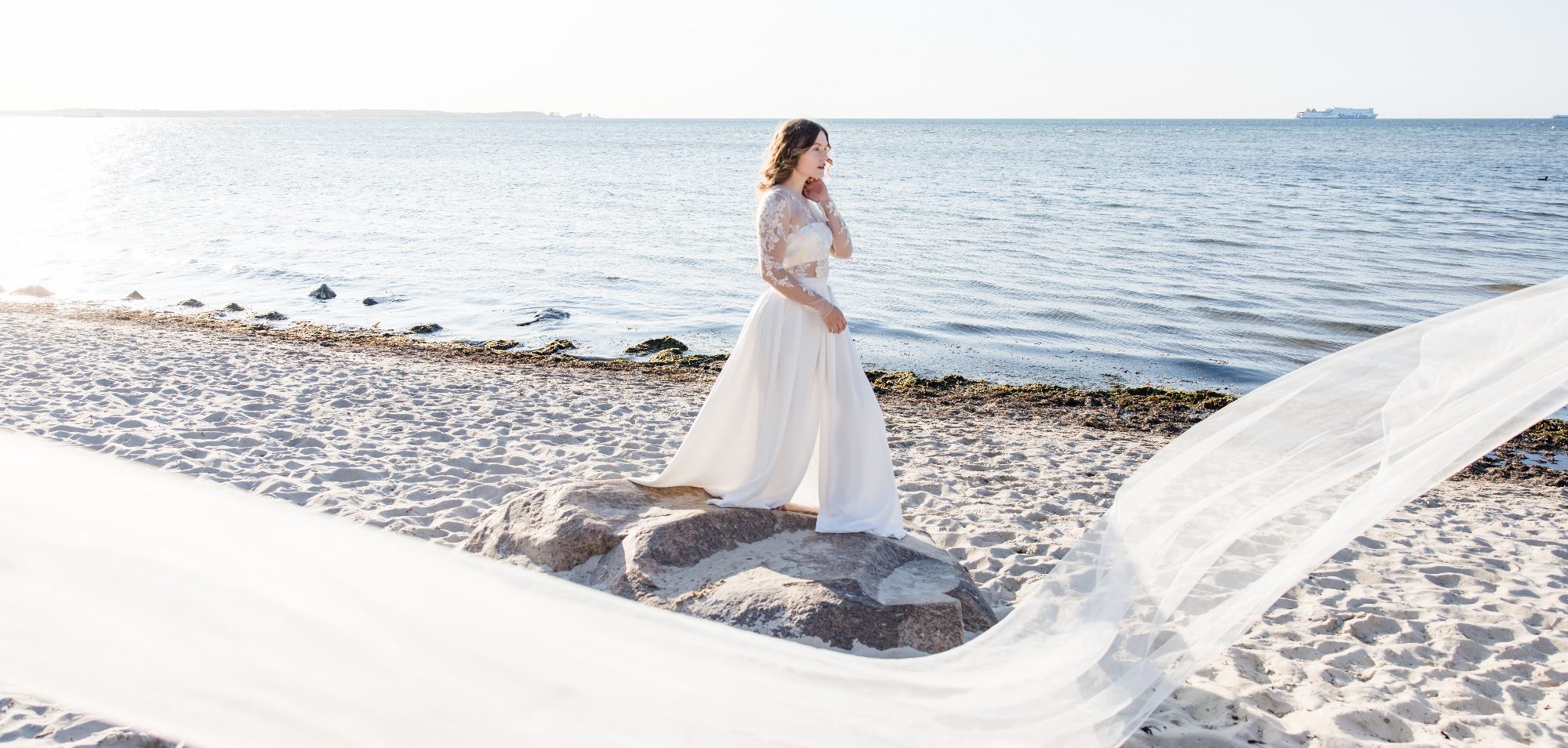 Braut in einem Zweiteiler als Brautoutfit. Ein Crepe Rock mit Beinschlitz weht im Wind am Strand. Ein transparentes Blumenoberteil strahlt.