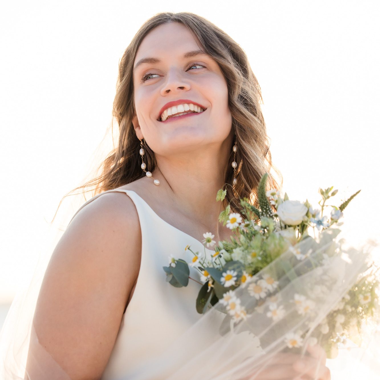Strahlende Braut mit Perlenohrringen und einem Wildblumen-Brautstrauß. Ein Fit&Flare Brautkleid - perfekt für deine Strandhochzeit.