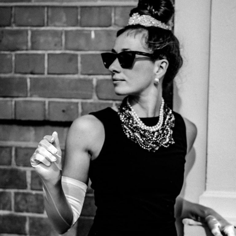 Eine  Frau steht in einem schwarzen Kleid mit viel Schmuck und weißen Handschuhen vor einer Wand.