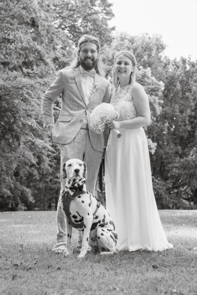 Braut strahlt mit Ehemann im Arm und Hund sitzen davor.
