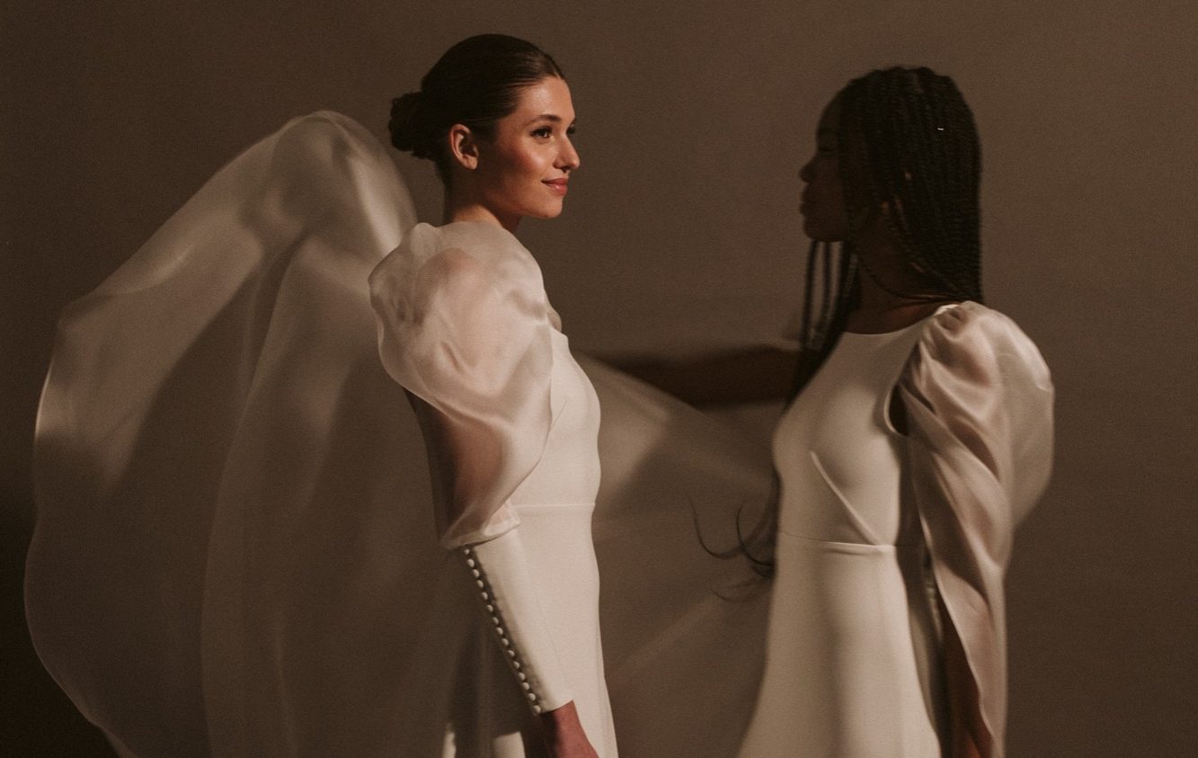 Braut steht am Strand vor der Wasserkante und ein Schleier weht vor ihr im Wind.