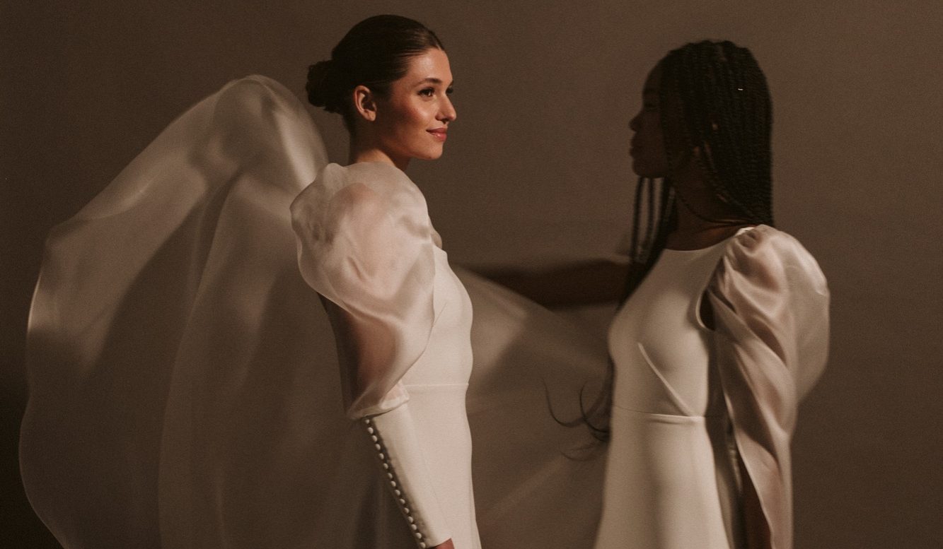Braut steht am Strand vor der Wasserkante und ein Schleier weht vor ihr im Wind.