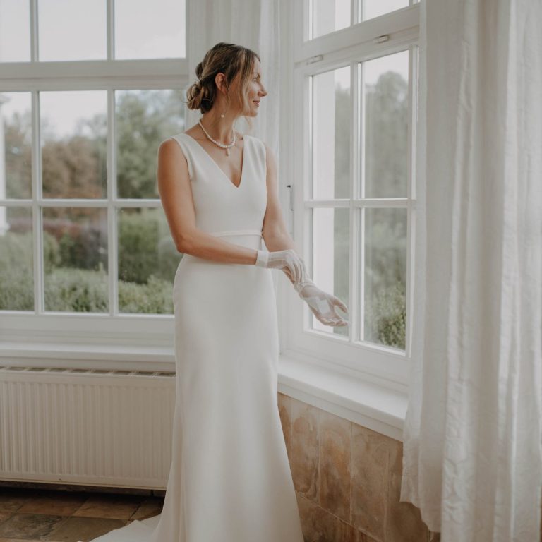 Fit&Flaire Brautkleid. Braut schaut aus dem Fenster auf die Hochzeit im Garten. Zarte Handschuhe und ein Clean Bridal Look.