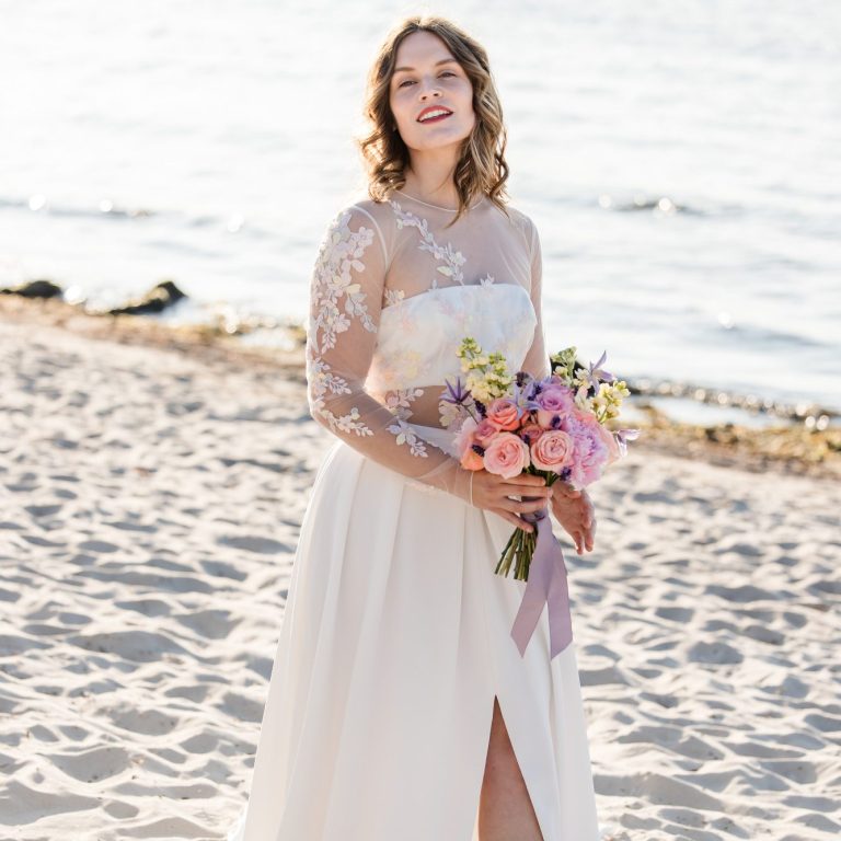 Braut am Ostseestrand bei einer Strandhochzeit. Ein Oberteil mit pastellfarbigen Blumen und einen Brautstrauß in der Hand.