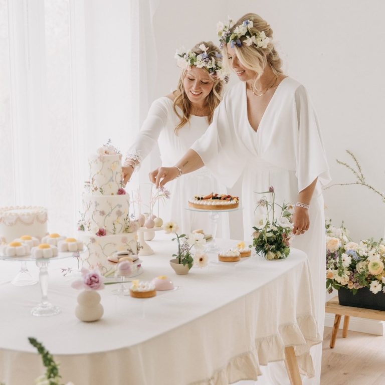 Brautmode mit Jumpern. Bräute stehen vor der Candy-Bar mit Hochzeitstorte. Beide tragen Blumenkränze als Brautaccessoire.